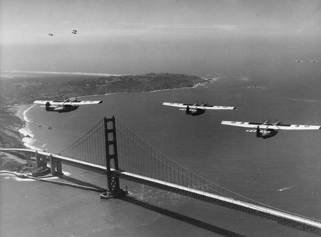 Three large aircraft fly above a suspension bridge, with smaller aircraft in the distance and a curved shoreline beyond.