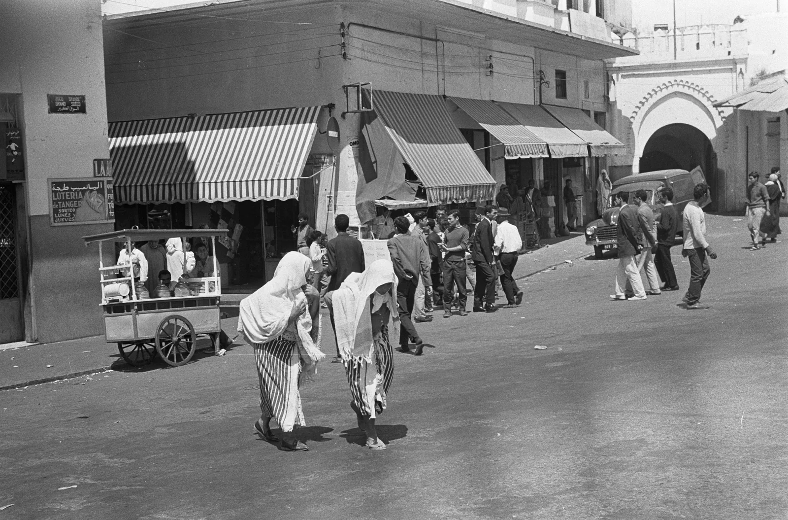 An archival street scene with shop awnings, a market cart, and a stone gateway
